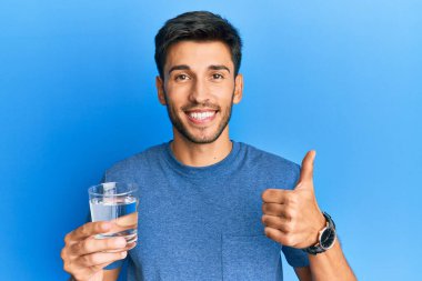 Genç yakışıklı adam bir bardak su içiyor mutlu ve pozitif gülümsüyor, başparmağıyla mükemmel ve onay işareti yapıyor. 