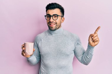 Genç İspanyol adam gözlük takıyor ve bir fincan kahve içiyor. Gülümsüyor. Elleri ve parmakları kenarda mutlu bir şekilde işaret ediyor. 