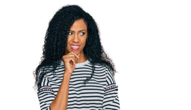 Orta yaşlı Afro-Amerikalı kadın. Elleri çenesinde gündelik kıyafetler giyip soru sormayı düşünüyor. Düşünceli bir ifade. Düşünceli bir yüzle gülümsüyor. şüphe kavramı. 