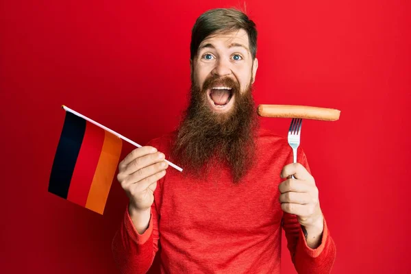 Redhead man with long beard holding fork with pork sausage and germany flag celebrating crazy and amazed for success with open eyes screaming excited.
