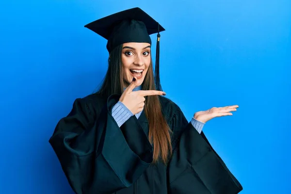 美しいブルネット若い女性が卒業キャップと式のローブを身に着けていると手で提示しながら カメラに驚いて笑顔と指で指して — ストック写真