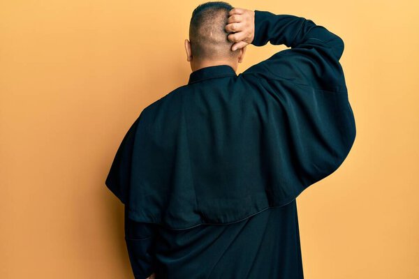 Young latin priest man standing over yellow background backwards thinking about doubt with hand on head