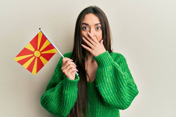Молодая латиноамериканка, держащая македонский флаг, прикрывающий рот рукой, шокирована и боится ошибиться. Удивительное выражение 