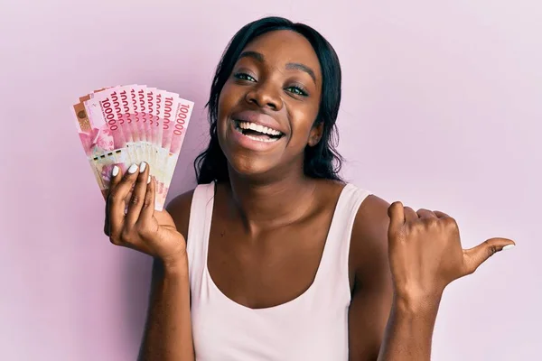 Young African American Woman Holding Indonesian Rupiah Banknotes Pointing Thumb — Stock fotografie