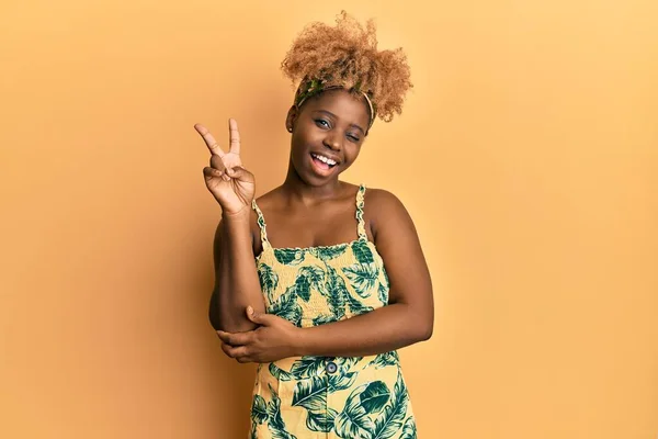 夏のドレスを着てアフロの髪を持つ若いアフリカの女性は 勝利の兆候を行うカメラでの幸せな顔のウィンクで笑顔 — ストック写真