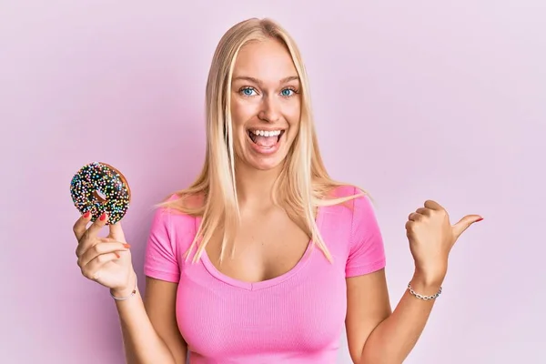 Jovem Loira Segurando Donut Apontando Polegar Até Lado Sorrindo Feliz — Fotografia de Stock