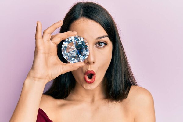 Молодая латиноамериканка, держащая бриллиантовый камень на глазу испуганная и изумленная открытым ртом для удивления, неверного лица 