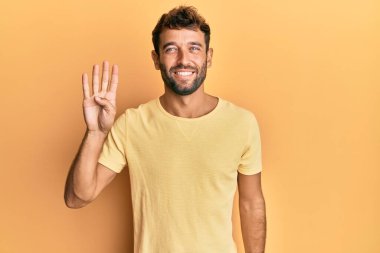 Sarı arka planda sarı tişört giyen sakallı yakışıklı bir adam dört numarayı gösterirken kendine güvenen ve mutlu bir şekilde gülümsüyor.. 