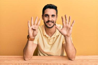 Latin asıllı genç adam, gündelik giysiler içinde masada oturuyor ve dokuz numaralı parmaklarını göstererek gülümsüyor ve mutlu.. 