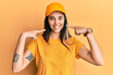 Teslimat üniforması giyen, şapkalı, neşeli gülümseyen, parmaklarıyla ve ağzıyla işaret eden genç İspanyol kadın. diş sağlığı kavramı. 