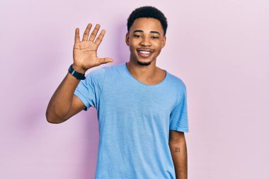 Genç Afro-Amerikan adam günlük mavi tişört giyer ve parmaklarıyla 5 numarayı gösterirken kendinden emin ve mutlu bir şekilde gülümser.. 