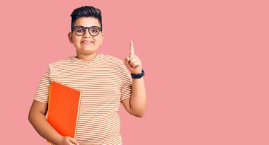 Küçük çocuk elinde kitapla gülümsüyor şaşkın şaşkın ve parmaklarıyla işaret ediyor ve kollarını kaldırıyor.. 