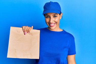 Elinde kağıt poşet tutan güzel esmer kadın pozitif ve mutlu görünüyor ve kendine güvenen bir gülümsemeyle dişlerini gösteriyor. 