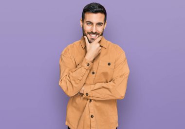 Gündelik kıyafetler giyen genç İspanyol adam kameraya kendinden emin bakıyor kollarını kavuşturup gülümsüyor ve çenesini kaldırıyor. Olumlu düşünmek. 