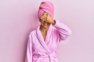 Orta yaşlı İspanyol kadın duş havlusu şapkası ve bornoz takıyor, gülümsüyor ve elleri yüzünde şaşkınlıktan gözleri kapatıyordu. kör kavram. 