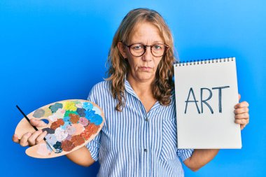 Boyacı paleti ve defter tutan orta yaşlı sarışın kadın sıkıntılı ve endişeli, sinirli ve korkmuş. hüzünlü ifade. 