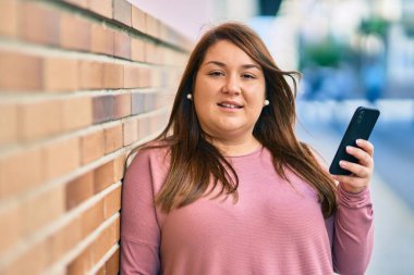 Genç ve cüsseli kadın şehirde akıllı telefon kullanarak mutlu bir şekilde gülümsüyor..