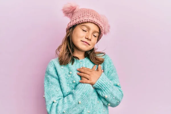 胸に手で笑みを浮かべてウールのセーターとかわいい冬の帽子を身に着けている小さな美しい女の子 目は顔に感謝のジェスチャーで閉じます 健康の概念 — ストック写真