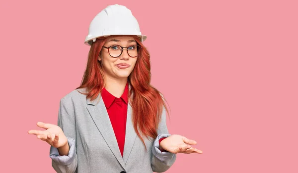 年轻的红头发女人戴着建筑师的硬礼帽 毫无头绪 满脸困惑 手臂和双手高举 怀疑概念 — 图库照片