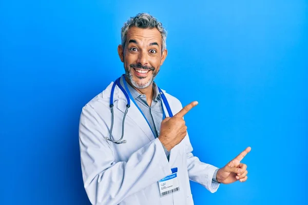 中年白发男子身穿医生制服 听诊器般的微笑着 看着摄像机 用双手和手指指向两侧 — 图库照片