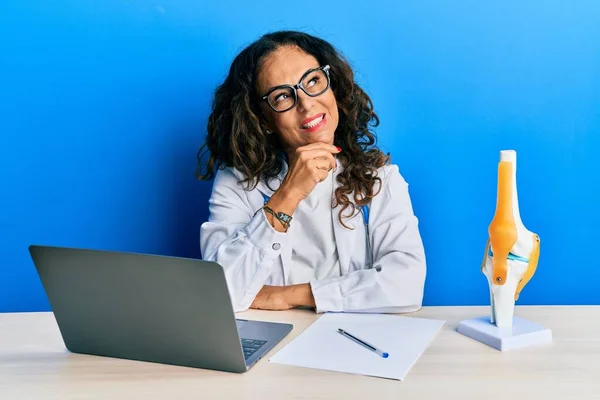 Beautiful middle age woman doctor at orthopedic clinic with hand on chin thinking about question, pensive expression. smiling with thoughtful face. doubt concept.