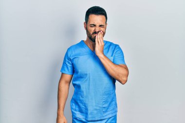 Sakallı, mavi erkek hemşire üniforması giyen yakışıklı İspanyol bir adam diş ağrısı ya da diş hastalığından dolayı ağızlarına acı dolu bir ifadeyle dokunuyor. dişçi 