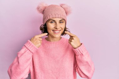 Şirin yün şapkalı, neşeli gülümseyen ve parmaklarıyla ve ağzıyla işaret eden genç esmer kadın. diş sağlığı kavramı. 