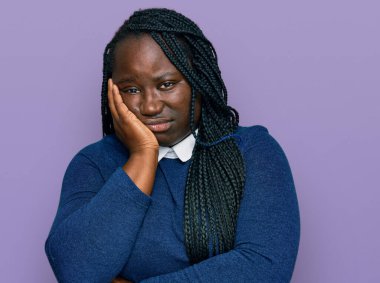 Saçları örgülü genç siyahi kadın kollarını kavuşturarak depresyondan yorgun ve sıkılmış göründüğünü düşünüyor.. 