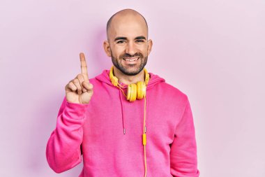 Spor kıyafetleri giyen ve kulaklık takan genç kel bir adam bir numarayı gösterip işaret ederken kendinden emin ve mutlu bir şekilde gülümsüyor.. 