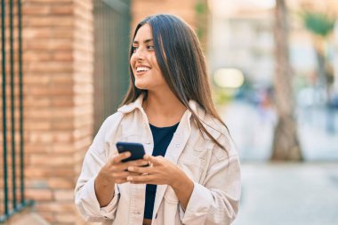Genç İspanyol kız şehirde akıllı telefon kullanarak mutlu bir şekilde gülümsüyor..
