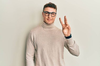 Balıkçı yaka kazak giyen İspanyol genç bir adam üç numaralı parmaklarıyla kendini beğenmiş ve mutlu gülümserken onu gösteriyor.. 