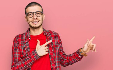 Latin asıllı, sıradan kıyafetler giyen, gülümseyen ve kameraya bakan genç bir adam. İki eliyle ve parmaklarıyla kenarı işaret ediyor.. 