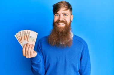 Genç İrlandalı kızıl saçlı adam elinde 5000 Güney Koreli para kazandı. Pozitif ve mutlu görünüyor ve gülümsüyor. Kendine güvenen bir gülümsemeyle dişlerini gösteriyor. 