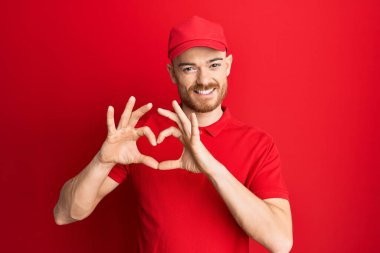 Genç kızıl saçlı adam teslimat üniforması ve şapkası takıyor. Aşkla gülümsüyor. Kalp sembolünü ve şeklini elleriyle gösteriyor. romantik kavram. 
