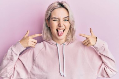 Genç sarışın kız parmaklarıyla ağzını işaret ediyor. Dilini çıkarıyor. Komik bir ifadeyle mutlu.. 