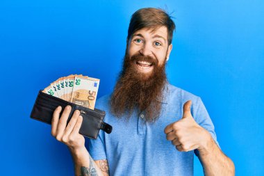 Uzun sakallı, elinde cüzdanı olan, mutlu ve olumlu gülümseyen, başparmağı mükemmel ve onay işareti yapan kızıl saçlı bir adam. 