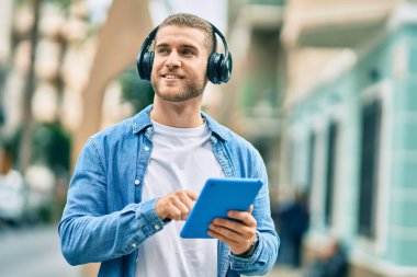 Genç beyaz adam şehirde kulaklık ve dokunmatik ped kullanarak mutlu bir şekilde gülümsüyor..