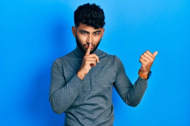 Balıkçı yaka kazak giyen sakallı bir Arap sessiz olmak istiyor. Parmakları dudaklarında, elleri yanda. sessizlik ve gizli konsept. 