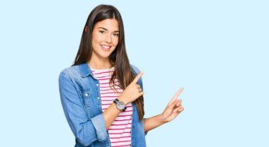 Genç esmer kadın, gündelik kıyafetler giyiyor gülümsüyor ve kameraya bakıyor. İki eliyle ve parmaklarıyla onu işaret ediyor.. 