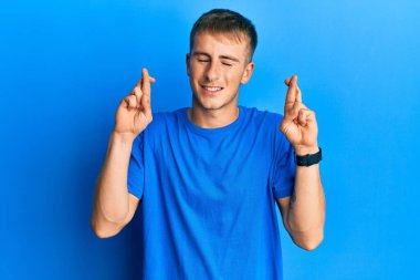 Sıradan mavi tişörtler giyen genç beyaz adam umut ve gözlerin kapalı bir şekilde gülümseyerek parmaklarını çapraz yaptı. Şans ve batıl inançlar. 