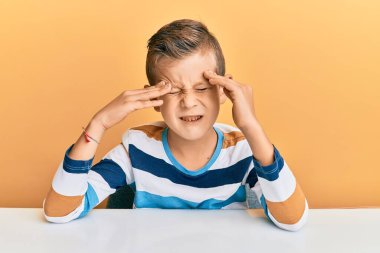 Kafkasyalı sevimli bir çocuk, günlük kıyafetler giymiş, eli başının üstünde masada oturuyor, stres yüzünden baş ağrısı çekiyor. migren ağrısı. 