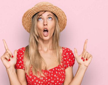 Yazlık şapkalı güzel beyaz kadın şaşırmış ve şaşırmış bir şekilde yukarı bakıyor ve parmaklarıyla işaret ediyor ve kollarını kaldırıyor.. 
