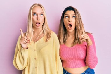 Pembe arka planda yan yana duran iki arkadaş şaşkınlık ve şaşkınlık içinde yukarı bakıp, parmaklarıyla işaret ederek ve kollarını kaldırarak. 