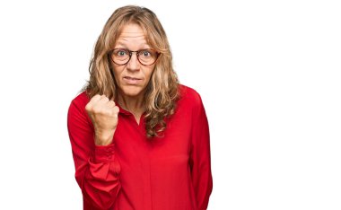 Orta yaşlı sarışın kadın, kırmızı arka plan üzerine gündelik tişört giyiyor kızgın ve kızgın yumruğunu kaldırıyor ve öfkeyle bağırıyor. öfke ve agresif konsept. 