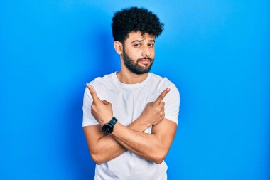 Sakallı, beyaz tişörtlü, iki tarafı da parmaklarıyla gösteren genç bir Arap, farklı yönlere bakıyor. 