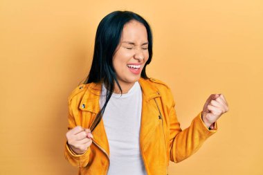 Sarı deri ceket giyen, burnu delinmiş güzel İspanyol bir kadın çok mutlu ve heyecanlı, kolları havada, gülümseyen ve başarı için çığlık atan, kazanan bir jest yapıyor. kutlama konsepti. 