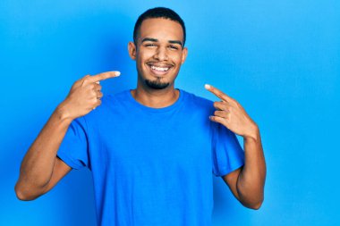 Gündelik mavi tişört giyen, gülümseyen, parmaklarıyla ve ağzıyla işaret eden Afro-Amerikan genç adam. diş sağlığı kavramı. 