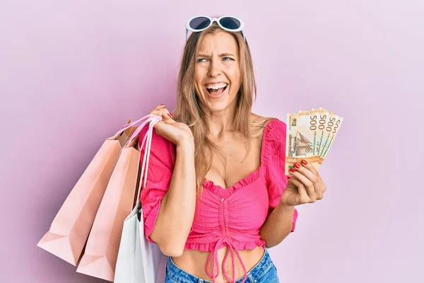 Beautiful caucasian woman holding shopping bags and 500 norwegian krone smiling and laughing hard out loud because funny crazy joke.