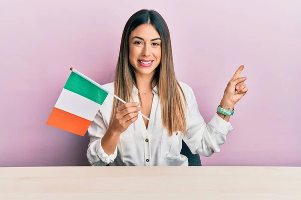 Mujer Hispana Joven Sosteniendo Bandera Irlandesa Sentada Mesa Sonriendo Feliz —  Fotos de Stock