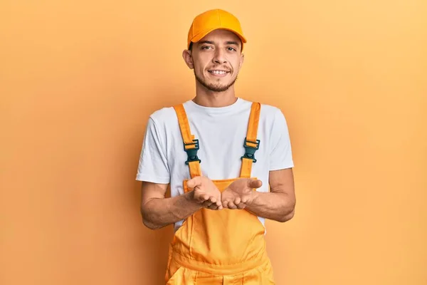 Spaanse Jongeman Draagt Klusjesman Uniform Glimlachend Met Handpalmen Samen Ontvangen — Stockfoto